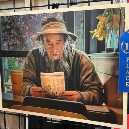 2024 Juried Art Show 1st place winner for Photography. Artwork called McDonald's Gandalf and depicts a man sitting at a table in a cafe with an iced coffee to one side, reading the newspaper, with a hat and coat that resemble the ones Gandalf wears